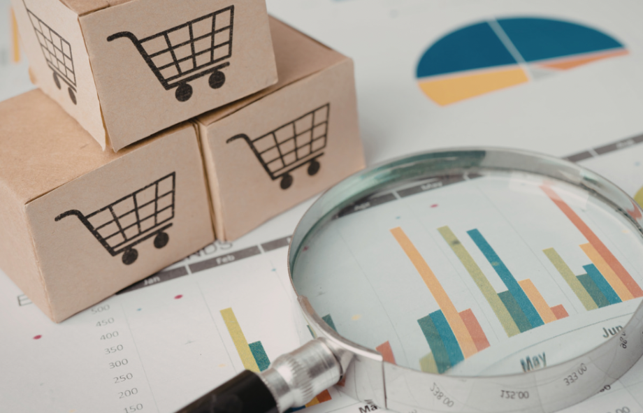 Miniature cardboard boxes with shopping cart icons stacked next to a magnifying glass placed over colorful business data charts.