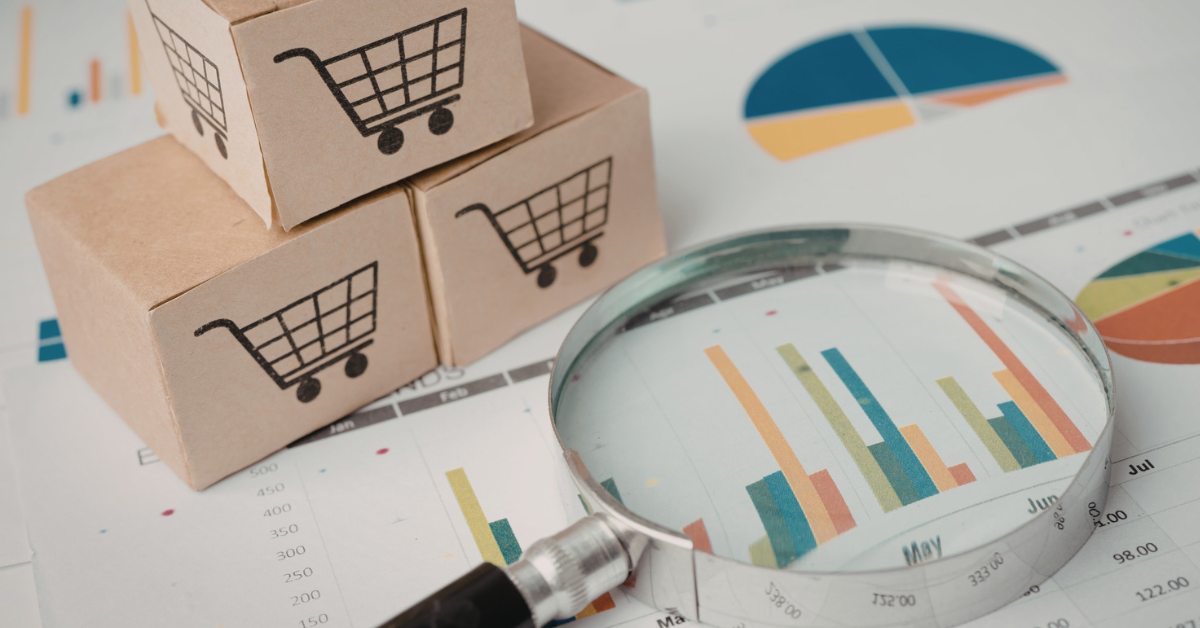Miniature cardboard boxes with shopping cart icons stacked next to a magnifying glass placed over colorful business data charts.
