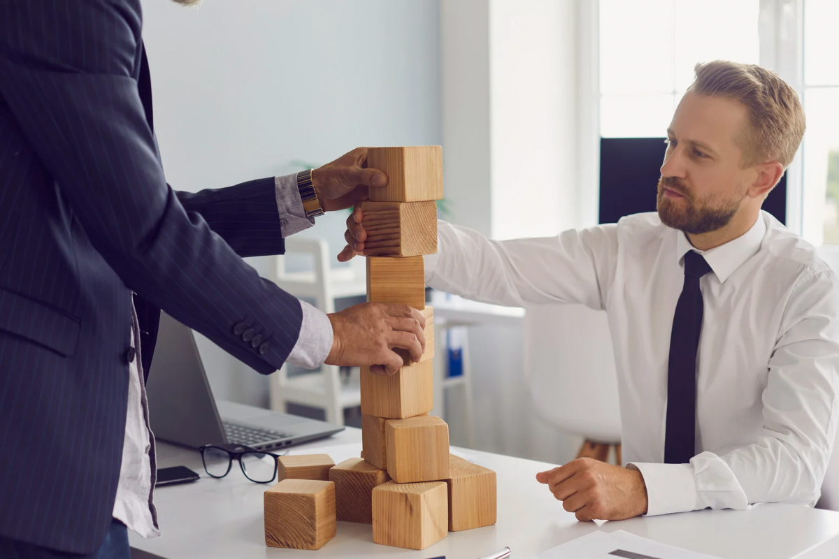 Two professionals stacking wooden blocks to represent the compounding, long-term stability of building owned brand real estate over renting ads.