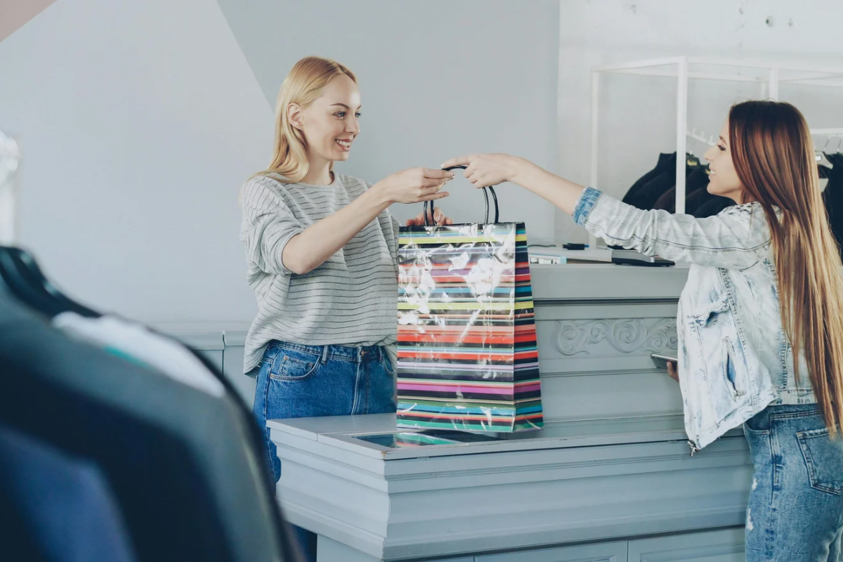 A retail worker handing a shopping bag to a customer across a checkout counter.