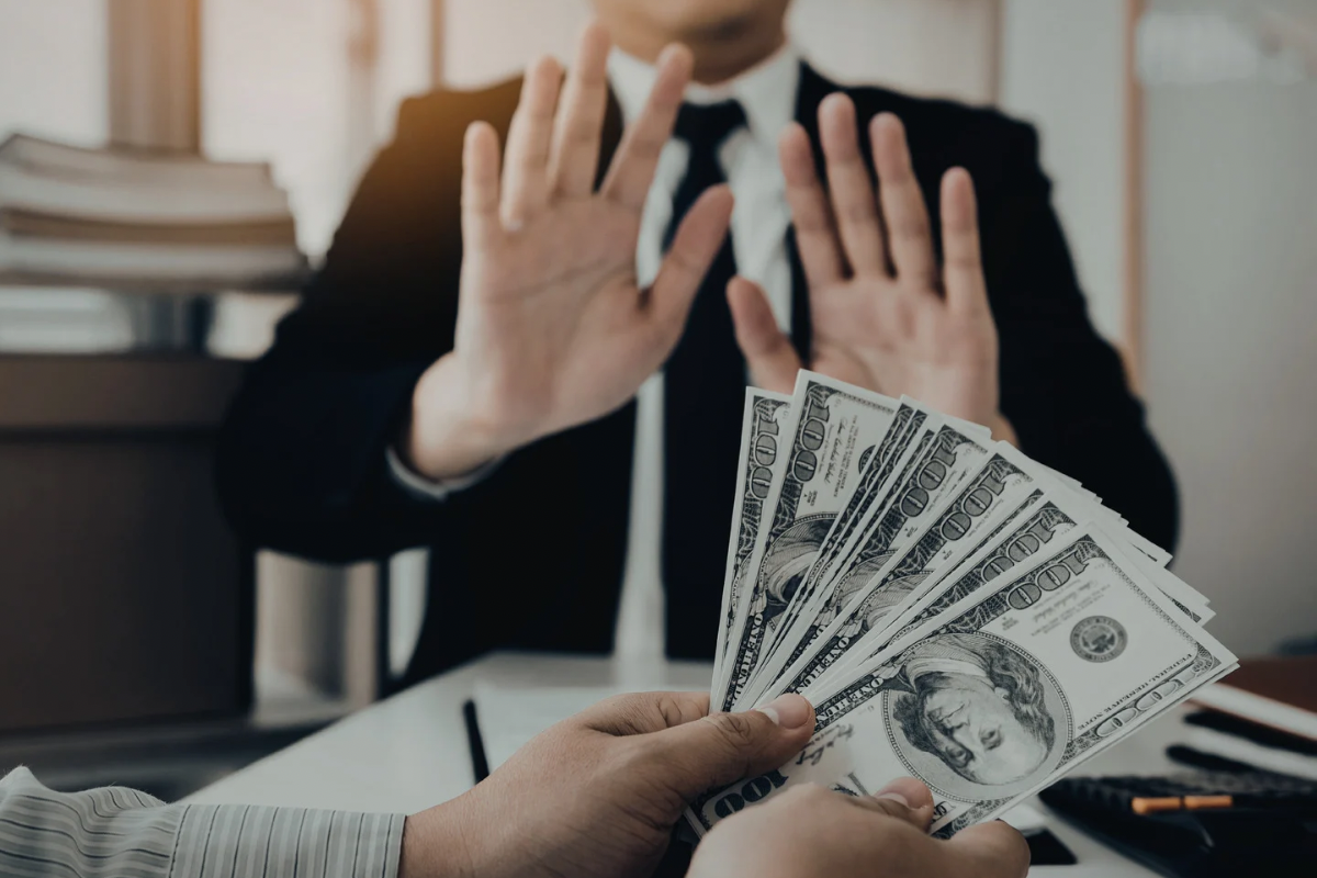 A businessman holding his hands up to politely refuse a fan of one-hundred dollar bills being handed to him.