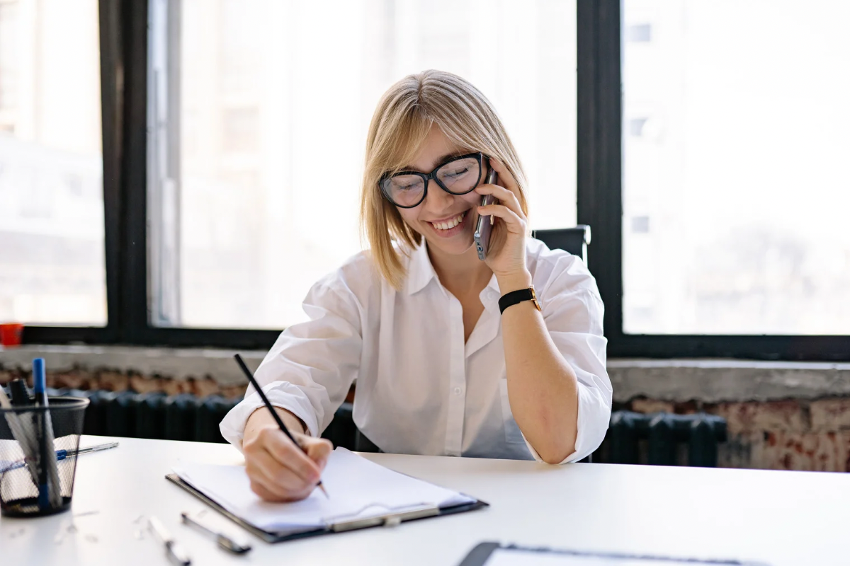 A woman on a phone call writing notes at a desk in an office.