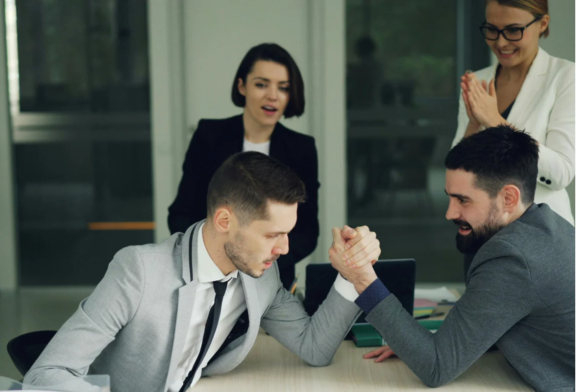 Two men in business suits arm-wrestling at an office desk while their colleagues watch.