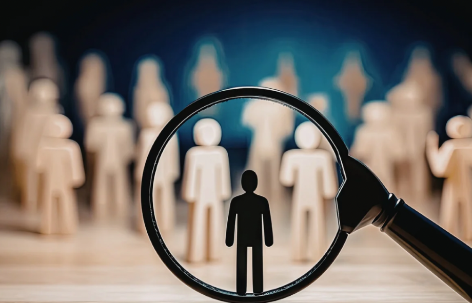 A magnifying glass focusing on a single black wooden figure standing out in a large crowd of identical beige figures.