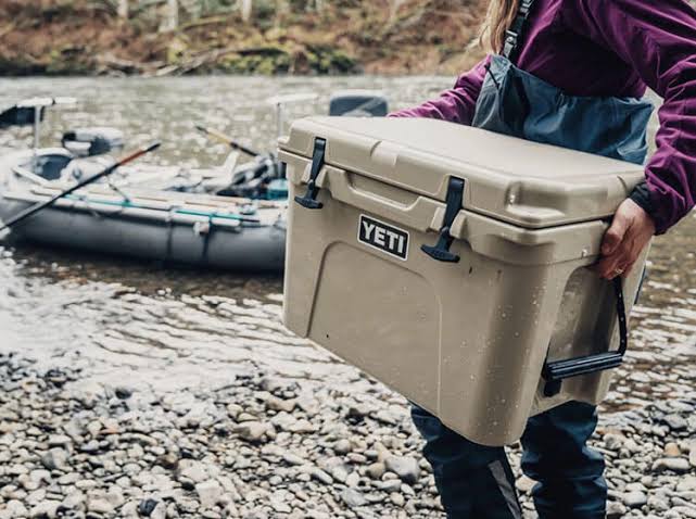 A person wearing outdoor wading gear carrying a premium tan YETI cooler along a rocky riverbank next to a boat.