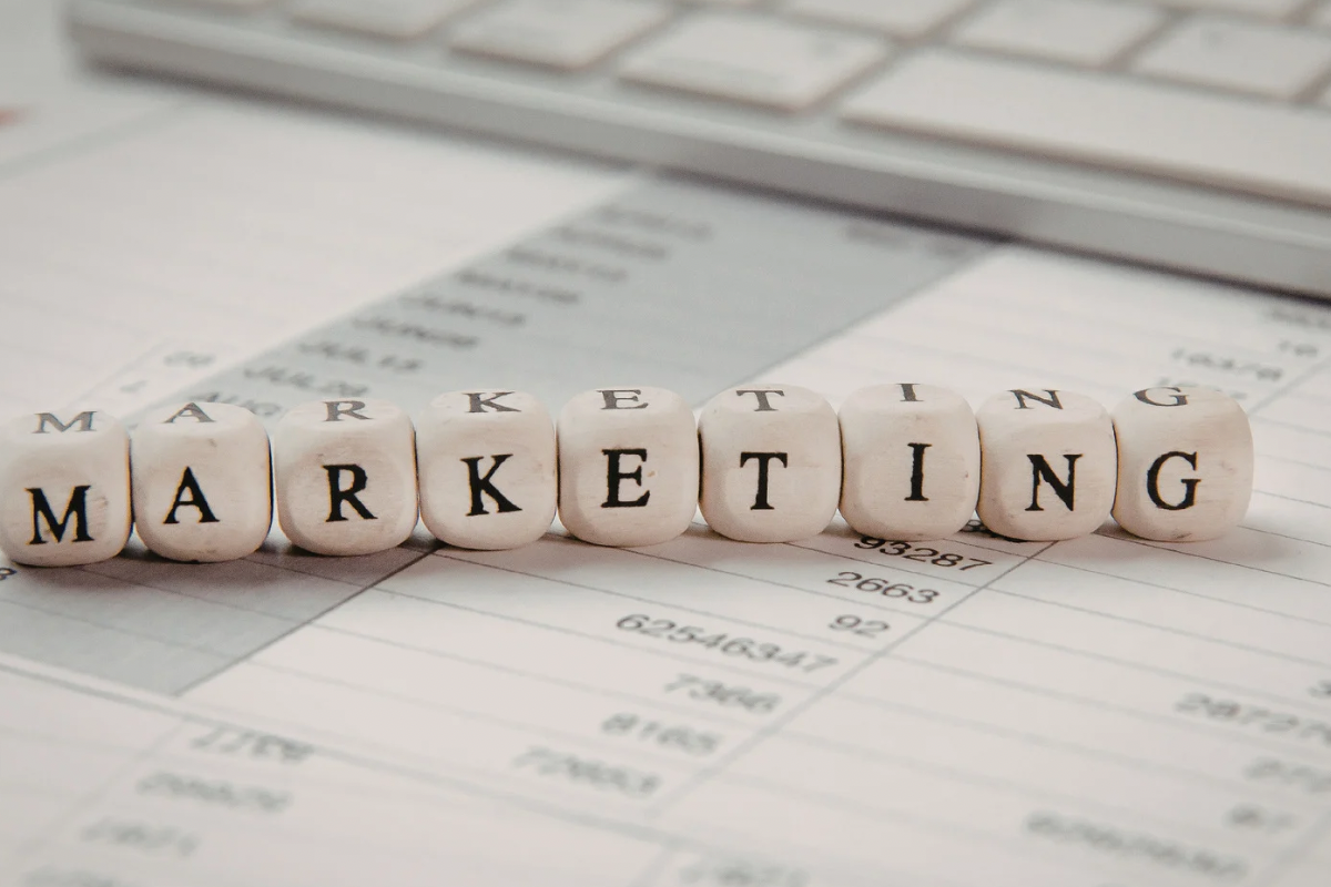 Wooden blocks spelling the word MARKETING sitting on top of data spreadsheets with a computer keyboard in the background.